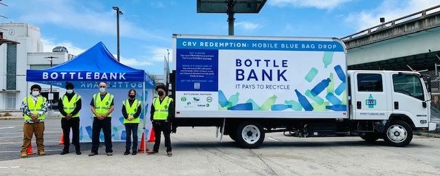 An industrial truck with the words "Bottle Bank" on the side is parked. Five staff members with bright yellow construction vests are standing next to the truck towards the back. 