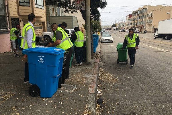 People in yellow vests pick up trash and wheel out trash and recycling bins on the curb