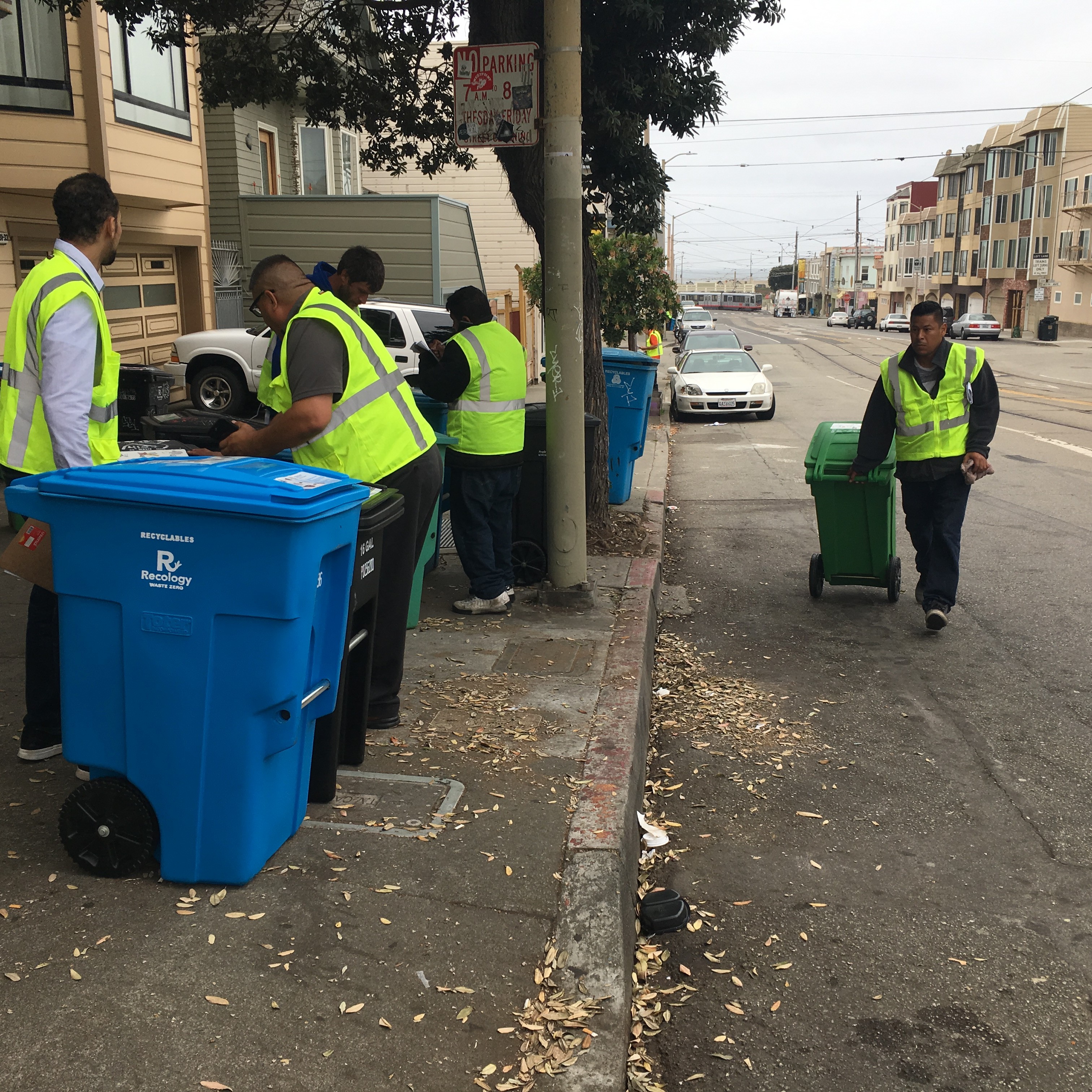 People in yellow vests pick up trash and wheel out trash and recycling bins on the curb