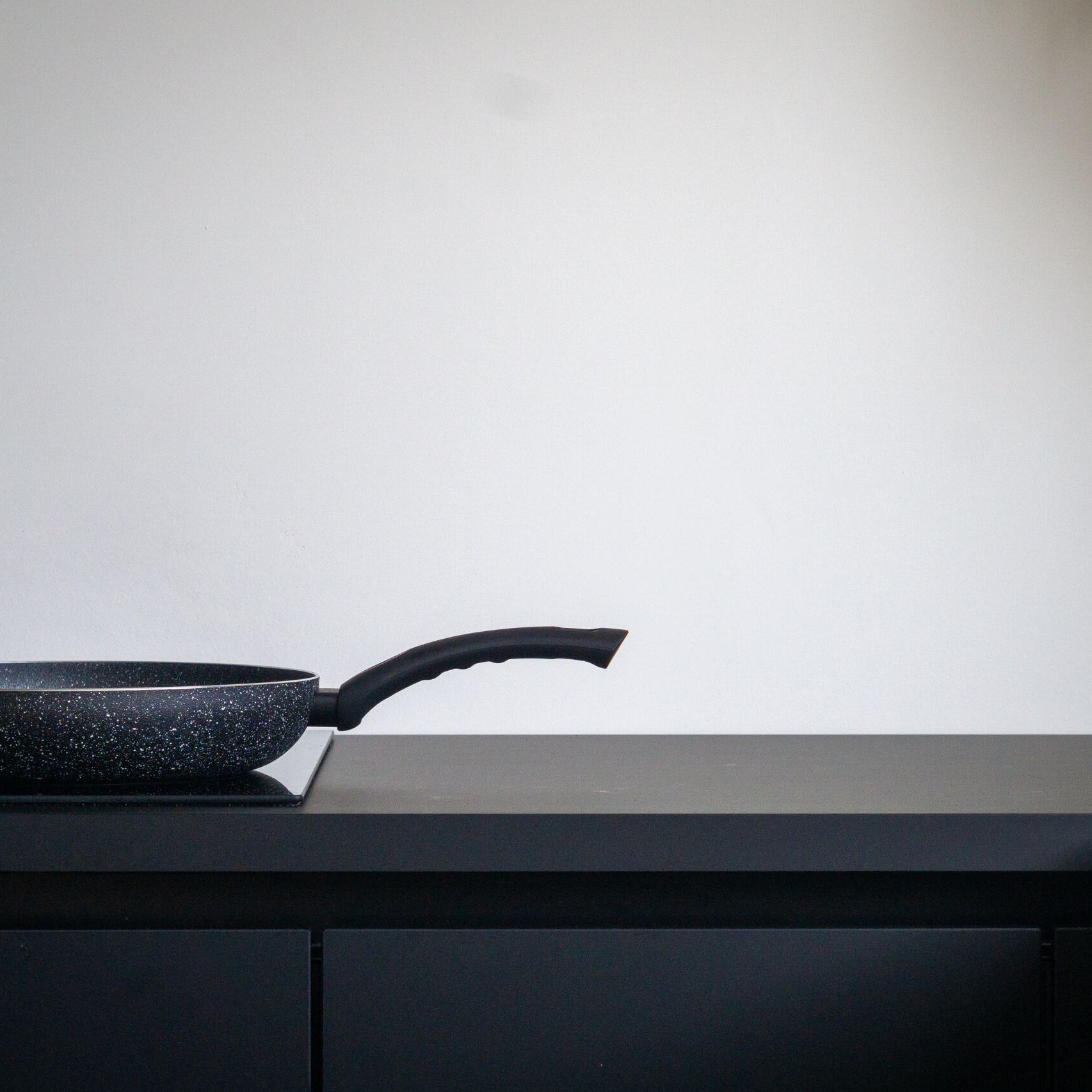 The image shows a black frying pan with a speckled pattern, placed on a black stovetop or countertop. The pan has a long, ergonomic handle. The background is a plain white wall, creating a minimalist and clean aesthetic. The scene is simple and uncluttered, focusing solely on the frying pan, which appears to be new and unused. The overall atmosphere is modern and sleek, with a strong emphasis on the contrasting colors of black and white.