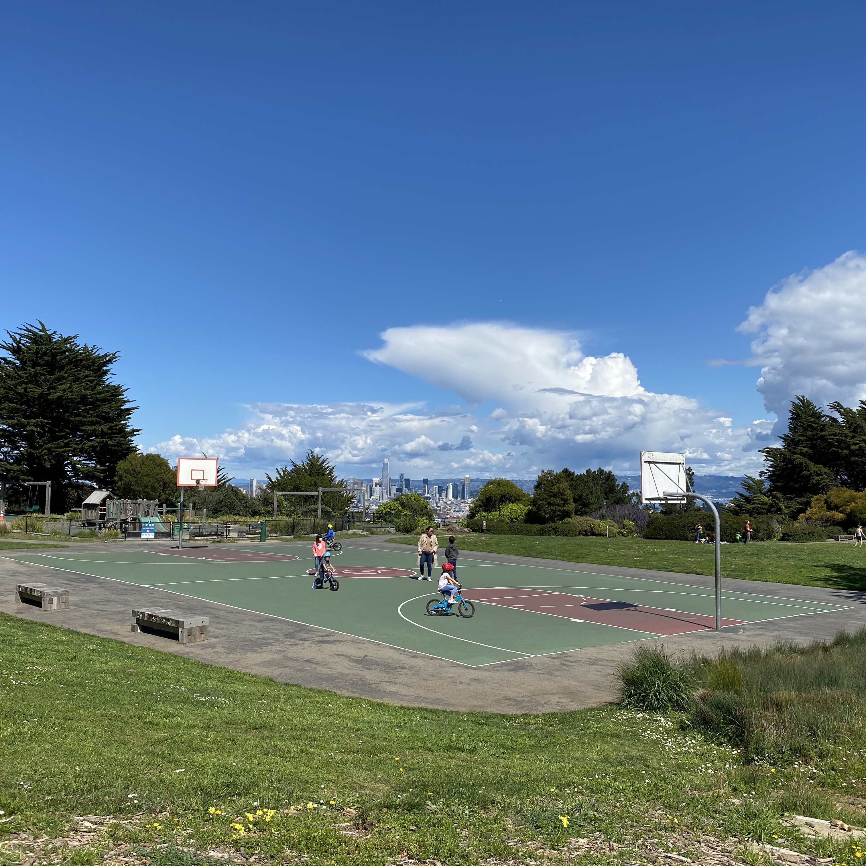 A sunny outdoor basketball court with children riding bicycles on it. The court is surrounded by green grass and benches. In the background, there are tall trees, a playground, and a clear view of the city skyline under a bright blue sky with scattered clouds.